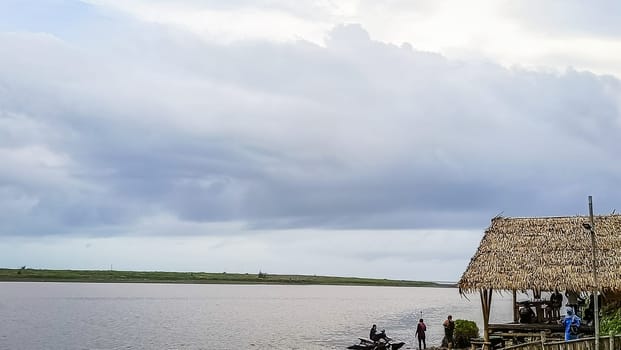 A beach with a small hut and a boat in the water. The sky is cloudy and the water is calm