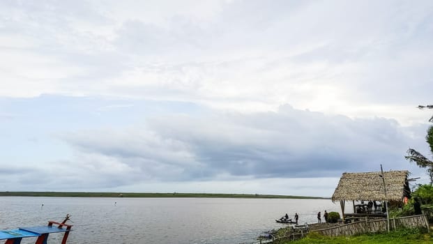 A beach with a small hut and a boat in the water. The sky is cloudy and the water is calm