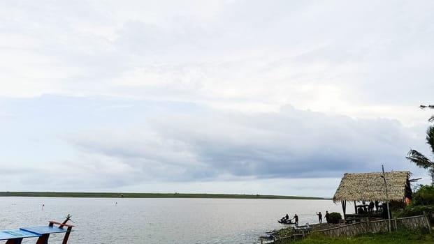 A beach with a small hut and a boat in the water. The sky is cloudy and the water is calm