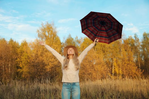 Happy woman in autumn park with umbrella raising her hands up, enjoying warm weather, trees with yellow leaves, nature forest background