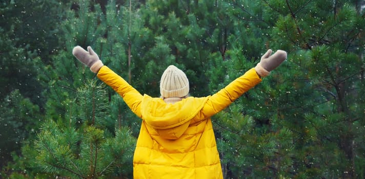Back view, winter day, happy young woman enjoys snow, warm weather against Christmas tree, joyful girl in hat, yellow jacket raising her hands up in snowy green forest with snowflakes