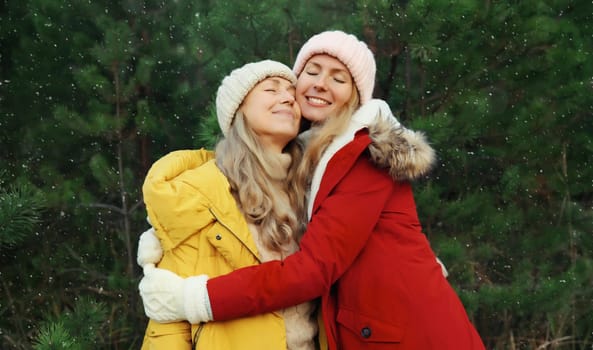 Happy smiling mature mother and adult daughter together hugging in winter forest against Christmas tree with snow, warm feelings, girlfriends in jacket, hat, nature background