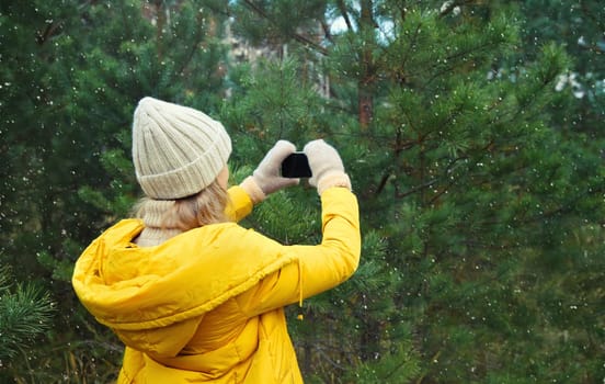 Young woman taking picture on phone Christmas tree in winter park, girl with smartphone outdoors, wearing yellow jacket, hat, standing in snowy forest with snowflakes, nature background