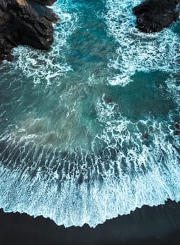A drone view of the waves during a storm in the Atlantic Ocean, a beach with black basalt sand and rocks on the islands, a scenic marine landscape without people with deep blue turquoise water.