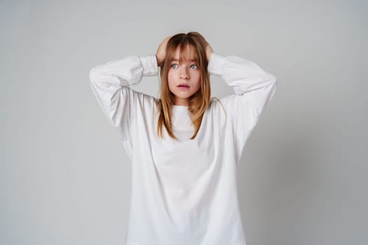 A teen girl in a loose white sweater looks surprised with her hands on her head against a plain gray backdrop close up