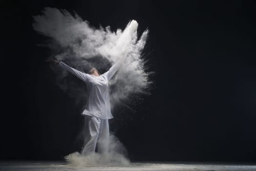 Man wearing white shirt and trousers jumping in white dust cloud in dark room full-length shot