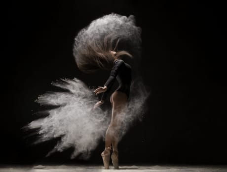 Woman in undrewear and pointe shoes jumping gracefully in a white dust cloudin dark room