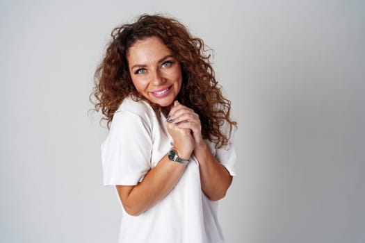 Cheerful woman with curly hair wearing a white shirt and jeans poses happily against a light gray background close up