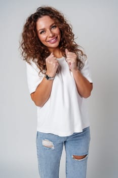 Cheerful woman with curly hair wearing a white shirt and jeans poses happily against a light gray background close up