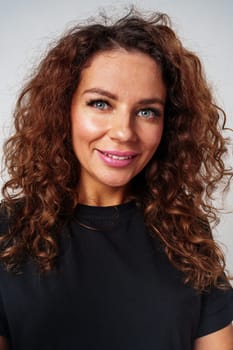 A woman with curly hair smiles brightly while wearing a black shirt against a neutral backdrop close up