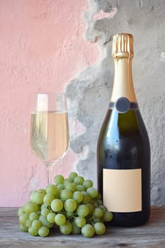 A bottle of champagne, a glass and a bunch of green grapes on a wooden table against a pink-gray textured plaster wall.