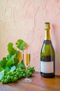 A bottle of champagne, a glass and a bunch of green grapes on a wooden table against a pink-gray textured plaster wall.