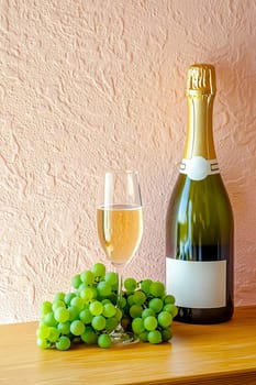 A bottle of champagne, a glass and a bunch of green grapes on a wooden table against a pink-gray textured plaster wall.