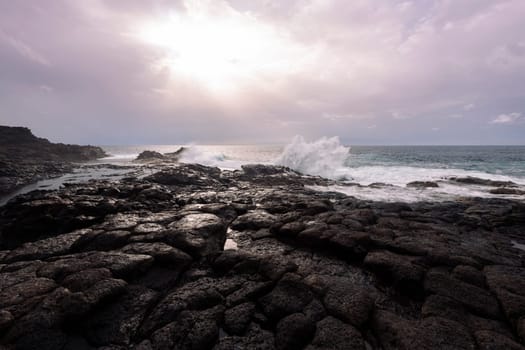 scenic view of foamy ocean against volcanic shore of lava black rocks and cloudy sky at sunset, concept of wild nature and marine landscape