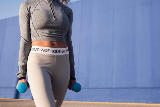 unrecognizable female athlete in sports clothes working out with dumbbells on urban roadway against blue wall in sunlight, concept of urban sport and healthy lifestyle, copy space for text