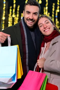 Smiling couple holding multiple colorful shopping bags, enjoying a festive evening out in the city. Dressed warmly for winter, they radiate joy and excitement, surrounded by holiday lights and a lively atmosphere.