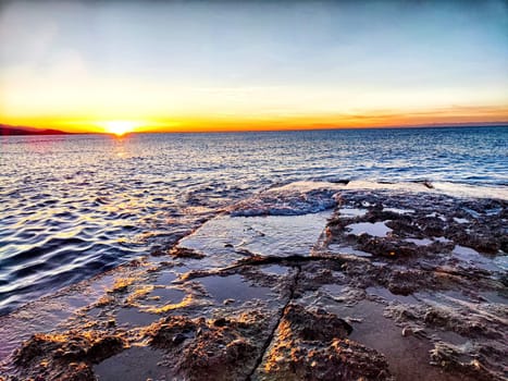 The sun sets on a tranquil shoreline, casting vibrant colors over the ocean while waves gently lap against the rocks.
