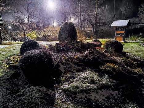 In a tranquil garden at night, snow gently covers bushes while soft lights cast an ethereal glow on the serene landscape.