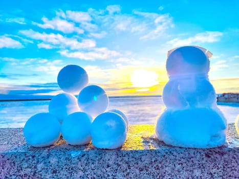 Snowballs and a snowman sit on a ledge during a colorful sunset over the water.