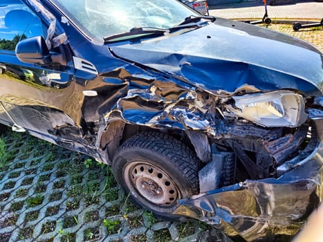 A black car shows significant damage to the front left side, parked near contemporary buildings in a sunny outdoor area