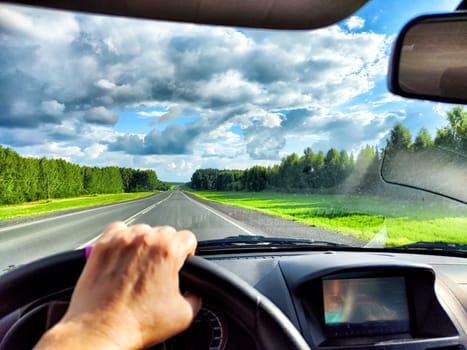 The driver steers along highway, with vibrant green fields and dramatic clouds stretching across the sky