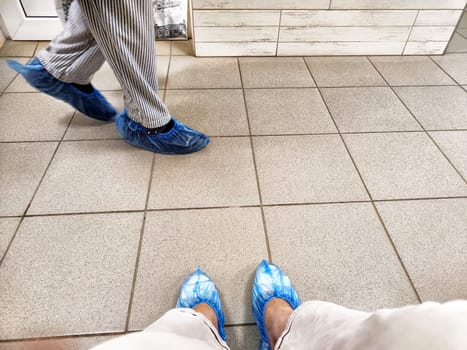A person is waiting in a doctor's clinic hallway, sitting with feet in blue shoe covers on tiled flooring