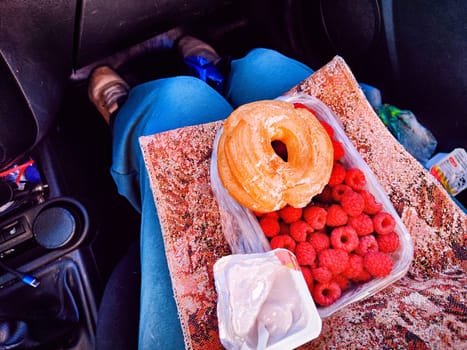 A delicious breakfast featuring a donut, fresh raspberries, and yogurt is set on a lap inside a car.