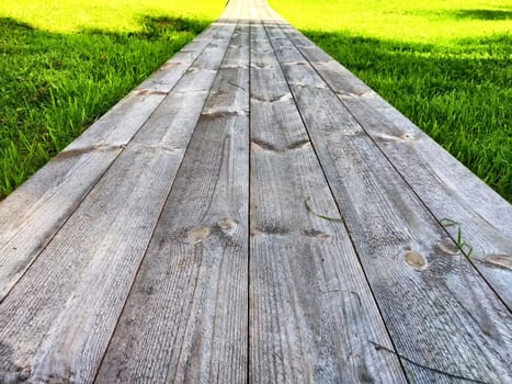 The rustic wooden pathway stretches forward, surrounded by vibrant green grass, inviting peaceful strolls in a tranquil garden.
