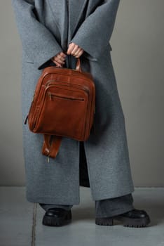 woman posing with a leather backpack wearing a warm gray coat and black boots.