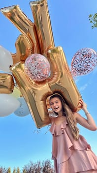 Smiling caucasian young girl model holding bunch of golden balloons smiling at camera. Blonde girl is wearing silk dress against blue sky. Birthday and celebration concept. Vertical