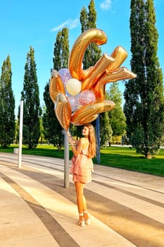Smiling caucasian young girl model holding bunch of golden balloons smiling at camera. Blonde girl is wearing silk dress against blue sky. Birthday and celebration concept. Vertical