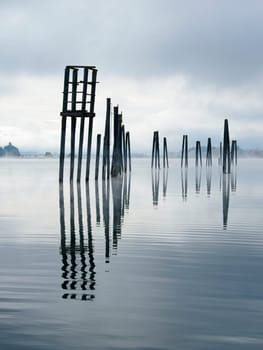 Tall wood pilings casting their reflections stand in the calm Pend Oreille River under a cloudy sky in eastern Washington.