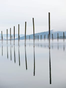 Tall wood pilings casting their reflections stand in the calm Pend Oreille River under a cloudy sky in eastern Washington.