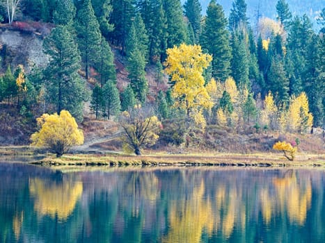 A seasonal landscape photo of fall colors from the trees reflecting off the calm Pend Oreille River in eastern Washington.