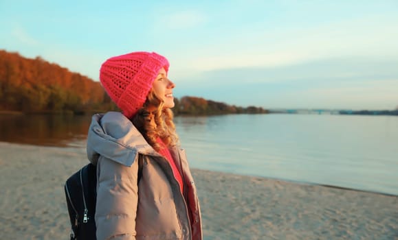 Travel, adventure, happy traveler young woman on the beach looking at amazing sunset, enjoying sunny landscape, warm weather, standing on sea coast, wear backpack, hat, jacket, nature background