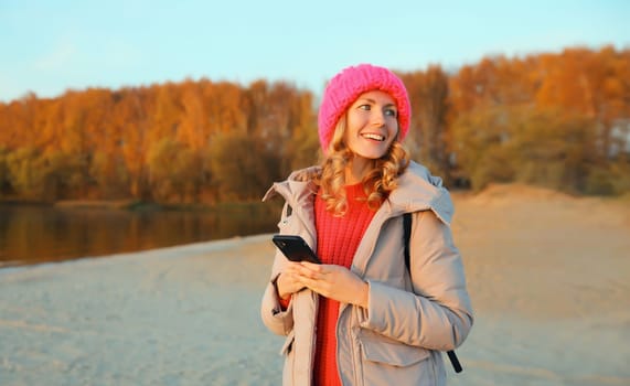 Travel, adventure, happy traveler young woman with phone on beach at sunset, girl holding smartphone standing on sea or river coast landscape, forest, wear backpack, hat, jacket, nature background