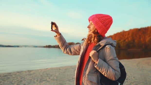 Travel, adventure, happy traveler young woman taking selfie with phone on the beach at sunset, girl using smartphone standing on sea or river coast with forest, trees, landscape, nature background