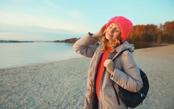 Travel, adventure, happy traveler young woman on the beach with beautiful sunset, enjoying sunny landscape, warm weather, standing on sea coast, wear backpack, hat, jacket, nature background