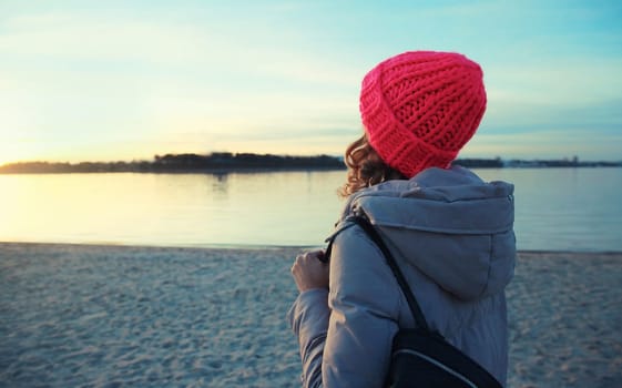 Travel, adventure, back view of traveler young woman on the beach looking at amazing sunset, enjoying landscape, standing on sea coast, wear backpack, hat, jacket, horizon nature background