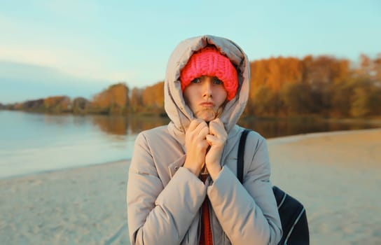 Woman is cold, freezing trying to warm up outdoors on the beach in autumn day, standing on sea coast, wearing warm jacket with hood, hat