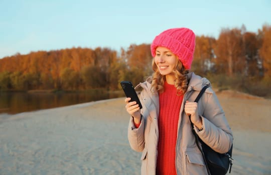 Travel, adventure, happy traveler young woman with phone on the beach at sunset, girl holding smartphone standing on sea or river coast with landscape wear backpack, hat, jacket, nature background