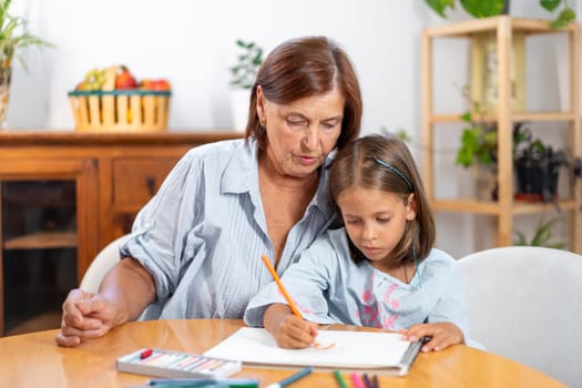 Senior woman watches fondly as her granddaughter draws with colored pencils in book, sharing nurturing and artistic moment together at home