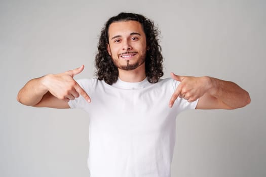 A young man with curly hair joyfully pointing down while wearing a plain white t-shirt against a neutral background close up