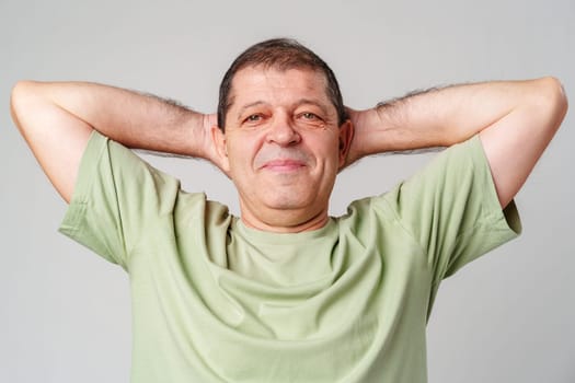 Happy older man in a green shirt smiling with arms behind his head in a bright studio setting close up