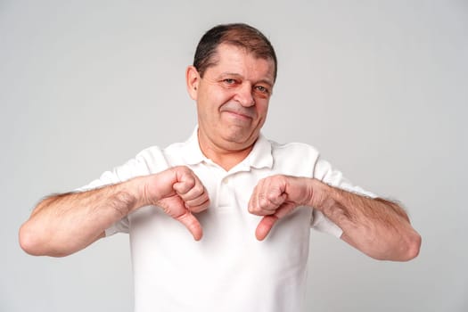 Older man expressing disapproval with thumbs down gesture while standing against a plain background close up