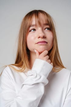 Young woman in a white long-sleeve shirt thoughtfully poses with a hand on her chin in a minimalistic indoor setting