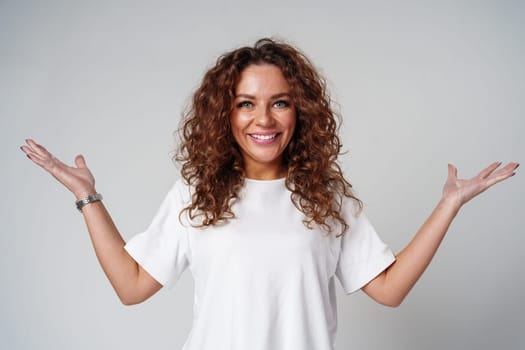 Cheerful woman with curly hair poses with open arms in a white shirt against a plain background close up