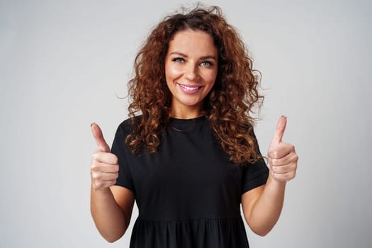 Cheerful young woman with curly hair giving a thumbs up while smiling against a neutral background close up