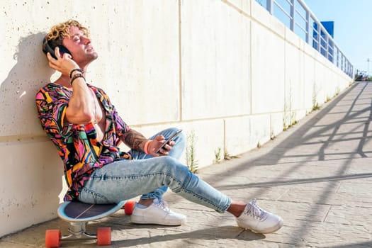 side view of dreamy male hipster sitting on skateboard on street and enjoying songs in headphones with closed eyes