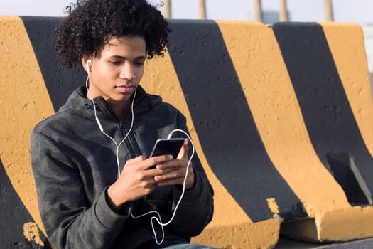 serious african american male teenager with earphones browsing on smartphone while sitting near striped wall on street, concept of technology of communication and young lifestyle